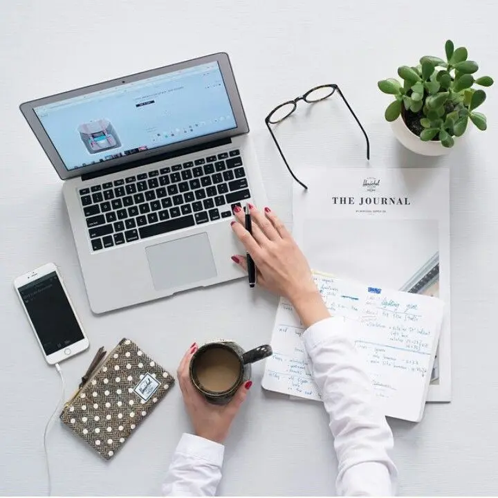 A person with red nails works at a laptop with a website open, surrounded by a journal, phone, glasses, a patterned wallet, a mug, and a plant, conveying a busy, organized workspace.